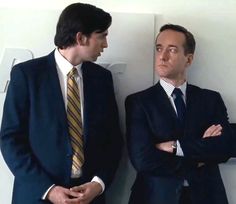 two men in suits standing next to each other with their arms crossed and looking at each other