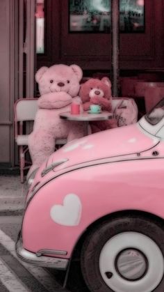 a pink vw bug parked in front of a building with two teddy bears sitting at a table