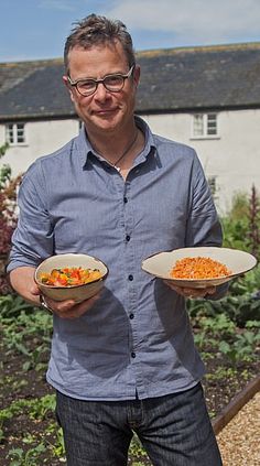 a man holding two plates of food in front of a house and garden with flowers