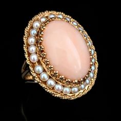 Step into a world of vintage charm with this mic last century substantial cocktail ring, crafted in gleaming 14k yellow gold. A smooth, large oval angelskin coral cabochon sits at the heart of this piece, framed by embellished gold prongs that elevate its presence like a beautiful frame. Surrounding the coral, a halo of lustrous pearls strung on gold wirework lends a touch of classic elegance. The ring's face is bordered by a twisted rope design, adding textural contrast and intricate detail. Supported by a robust triple split shank, this ring makes a bold yet sophisticated statement, perfectly capturing the 1940s dedication to craft and opulence Metals: 14k gold Markings: 14k hallmark Gemstones: coral - cabochon oval cut coral approximately 19x14.5mm, seed pearls Ring Size: 6.75 Total Wei High Luster Yellow Gold Oval Rings, High Luster Oval Ring In 14k Gold, High Luster Oval Yellow Gold Rings, High Luster 14k Gold Oval Rings, Classic Oval Ring With High Luster, Classic Oval Rings With High Luster, Classic Oval High Luster Ring, Luxury Round Jewelry For Vintage Events, High Luster Vintage Ring Jewelry