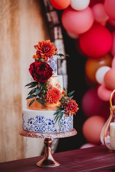 a multi - tiered cake with red flowers on top sits on a table in front of balloons