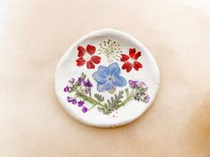 a white plate with red, blue and purple flowers painted on the front is sitting on a beige surface