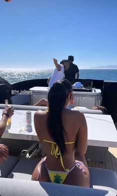 two women in bikinis sitting at a table on the back of a boat with people standing around