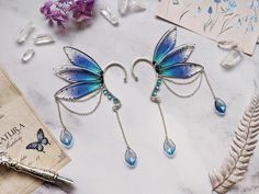 two blue butterfly earrings with dangling drops on them next to a book and some feathers