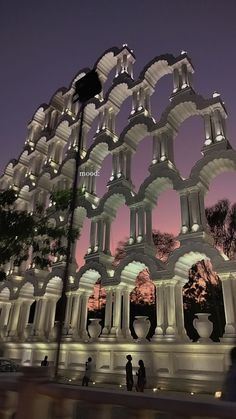 an artistic sculpture is lit up in the night sky with people walking around it at dusk