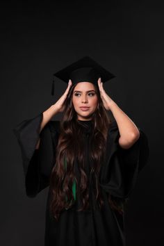 a woman wearing a graduation gown and holding her hands up to her head while standing in front of a black background