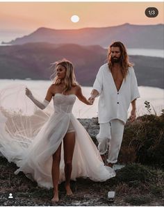 a man and woman holding hands while walking on top of a hill with the sun setting behind them