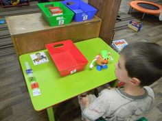 a young boy sitting at a green table in front of some toy bins on the floor