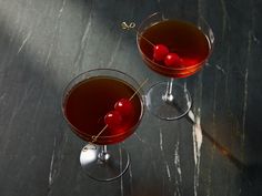 two wine glasses filled with red liquid and cherries on a black table top next to each other