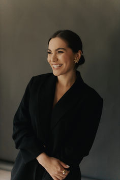 a smiling woman in a black suit and gold earrings, leaning against a gray wall