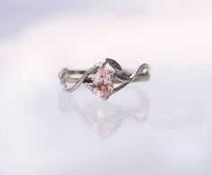 a white gold ring with an oval shaped morganite and leaves on the band, sitting on a reflective surface