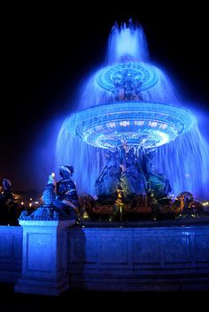 an image of a fountain with blue lights in the night sky and people around it