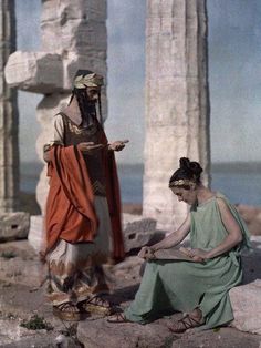 two women in ancient greek dress sitting on the ground next to some stone pillars and looking at their cell phones