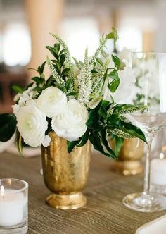 white flowers in a gold vase with greenery and candles on a table setting for a wedding reception