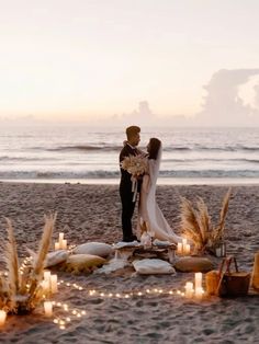 a man and woman standing on top of a sandy beach next to the ocean with candles