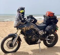 a motorcycle parked on top of a sandy beach next to the ocean with luggage on it's back