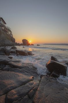 the sun is setting over the ocean with rocks in the foreground and houses on the other side