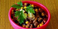 a red bowl filled with meat and vegetables on top of a wooden table next to a green leaf