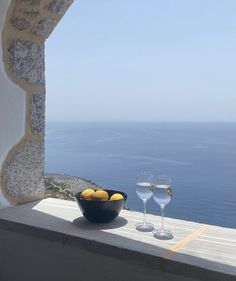 two wine glasses sitting on top of a window sill next to a bowl of fruit