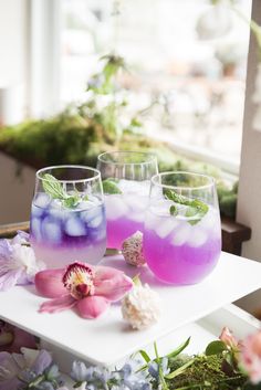 three glasses filled with different colored drinks on top of a white table next to flowers