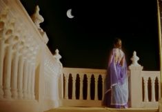 a woman in a blue sari standing on a balcony at night with the moon behind her