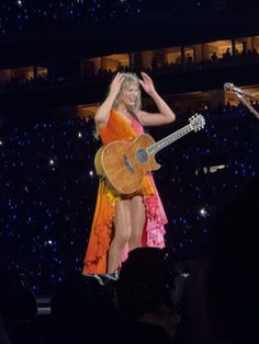 a woman in an orange dress is holding a guitar and standing on stage with her hands behind her head