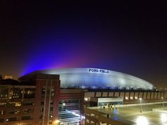 the stadium is lit up at night with bright lights on it's roof and people walking around