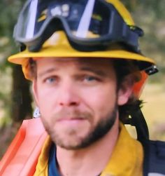 a man wearing a hard hat and goggles on his head with trees in the background