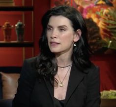 a woman with long black hair wearing a suit and necklace sitting in front of a red wall