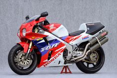 a red, white and blue motorcycle parked on top of a metal stand in front of a gray wall