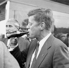 black and white photograph of man drinking from a glass in front of other people at an outdoor event