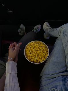 a person holding a bowl of food in their lap while sitting on the floor next to someone's feet