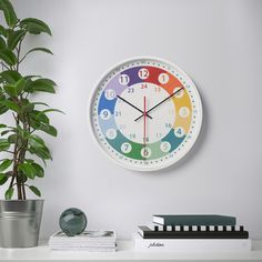 a white clock on the wall next to a potted plant and books in front of it