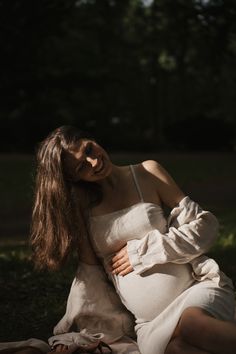 a pregnant woman sitting on the ground with her arms wrapped around her belly and looking off to the side