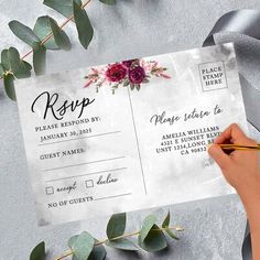 a person holding a pen and writing on a wedding rsp card with eucalyptus leaves around it