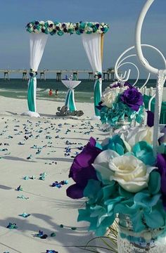 an outdoor wedding setup on the beach with purple and green flowers in vases, white drapes and blue petals