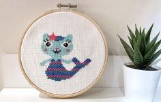 a cross - stitch cat is sitting on the shelf next to a potted plant