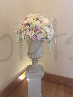 a white vase filled with lots of flowers on top of a wooden floor next to a wall