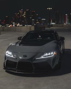 a gray sports car parked in front of a city skyline at night with the lights on