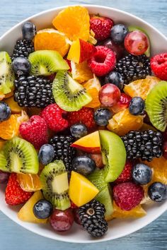 a white bowl filled with fresh fruit on top of a blue wooden table next to an orange, kiwi and raspberry