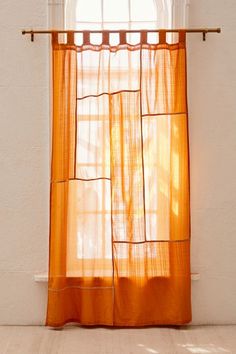 an orange sheer curtain hanging on a white wall next to a window with sunlight streaming through it