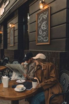 two people sitting at a table with coffee and newspaper in front of them on the sidewalk