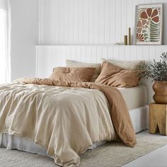 a bed with beige sheets and pillows in a white room next to a potted plant