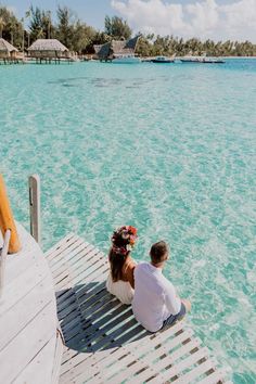 a man and woman sitting on a dock looking out at the water in front of some huts