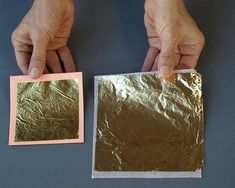 two hands are holding small square pieces of paper with gold foil on them, and one hand is holding a pink rectangle