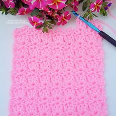 a crocheted pink square next to flowers and a blue crochet hook