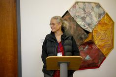 a woman standing at a podium in front of an art work on the wall behind her