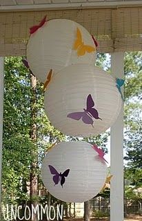 paper lanterns with butterflies hanging from them on a porch in front of some trees and bushes