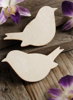 two wooden birds sitting on top of a piece of wood next to purple and white flowers