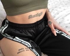 a woman with tattoos on her stomach sitting on a bed wearing black shorts and green shirt